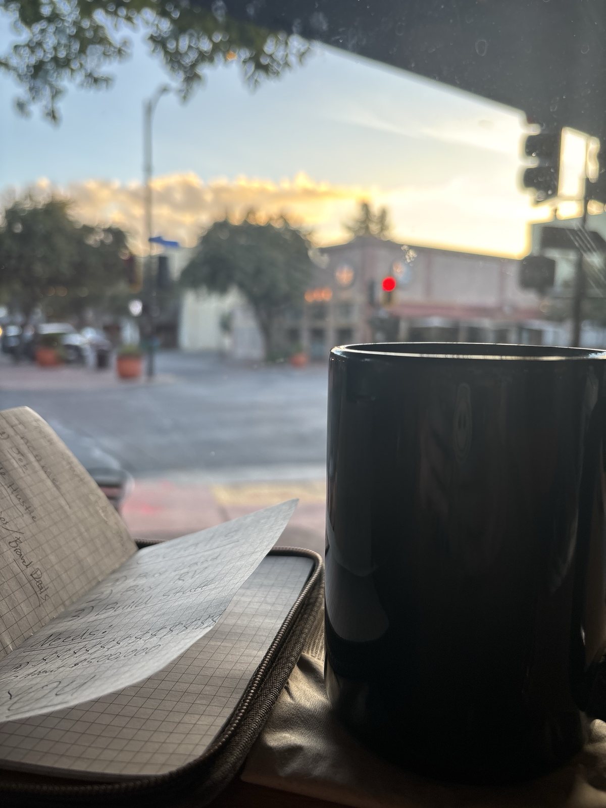 The notebook lies open on a windowsill next to a black coffee mug, with a street and a sunrise sky outside. The page lists Buy land, Buy RV, Build stuff and Needs.