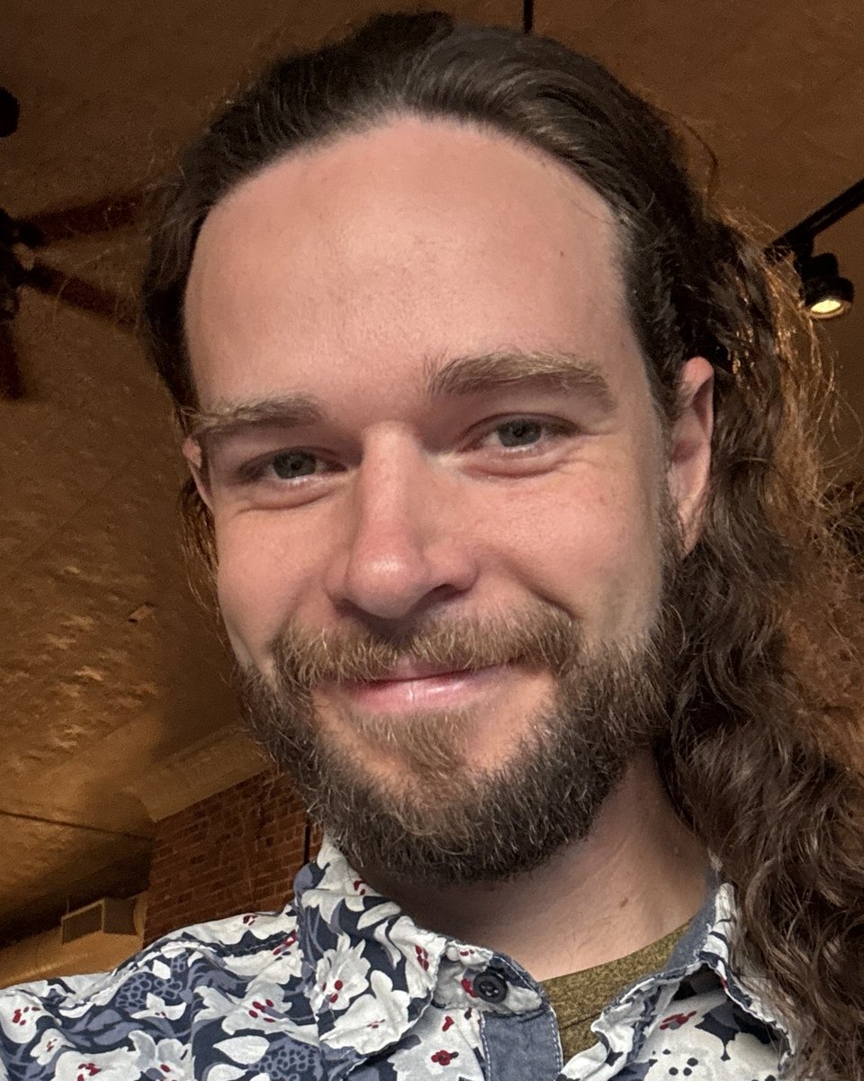 The artist smiling at the camera, long hair over one shoulder, in a blue and white flower-print shirt.